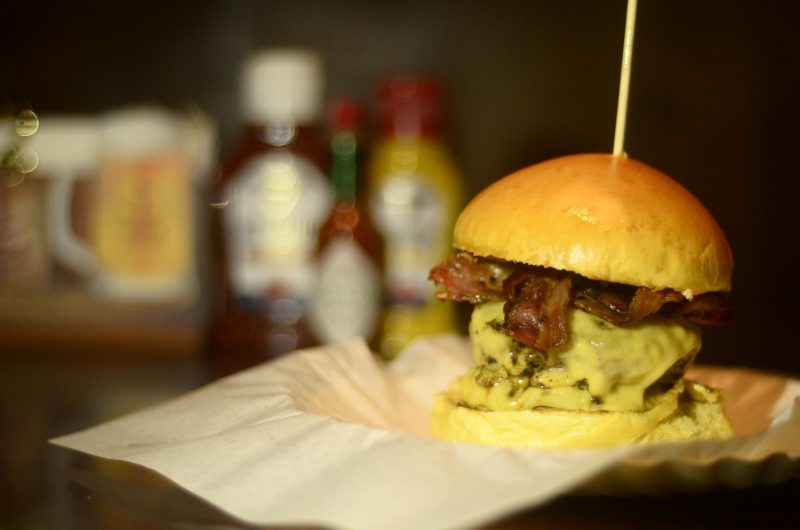 Hambúrguer com muito queijo e bacon em primeiro plano e molhos desfocados ao fundo