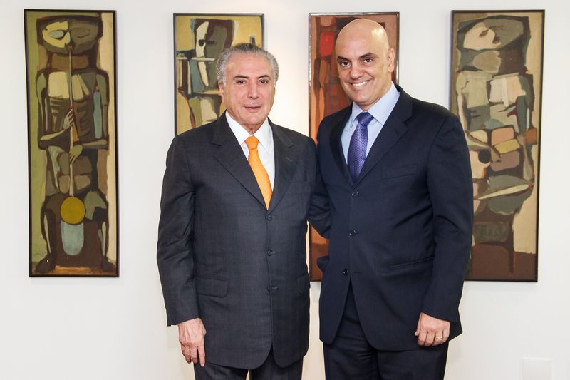 Dois homens brancos, de terno, posando para a foto em frente a parede branca com quadros 