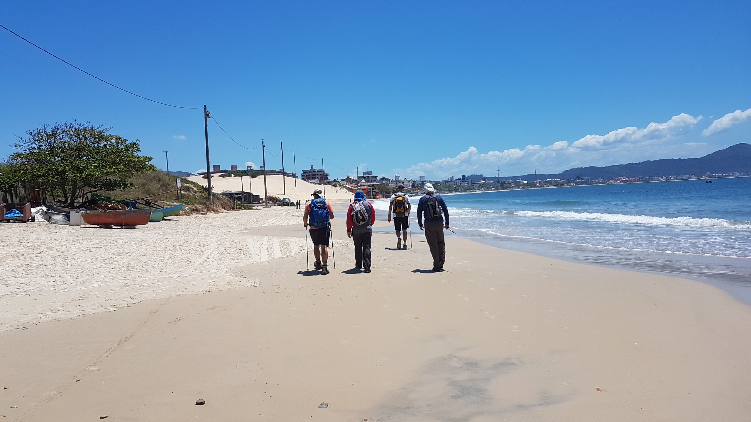 Foto de quatro homens de costas caminhando pela areia da Praia dos Ingleses. Eles seguram bastões de caminhadas e usam bonés. Do lado direito, o mar de águas azuis. Do lado esquerdo, alguns barcos na áreia e uma árvore. Ao fundo, dunas de areia e morros verdes. 