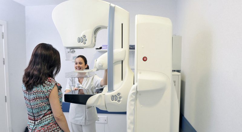 Enfermeira com roupa branca atrás de máquina de mamografia e paciente aparece de frente para ela mas de costas para a foto usando uma roupa com estampa colorida