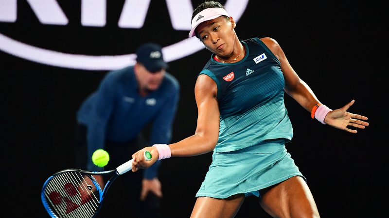 Naomi Osaka com roupa azul e batendo na bola com raquete