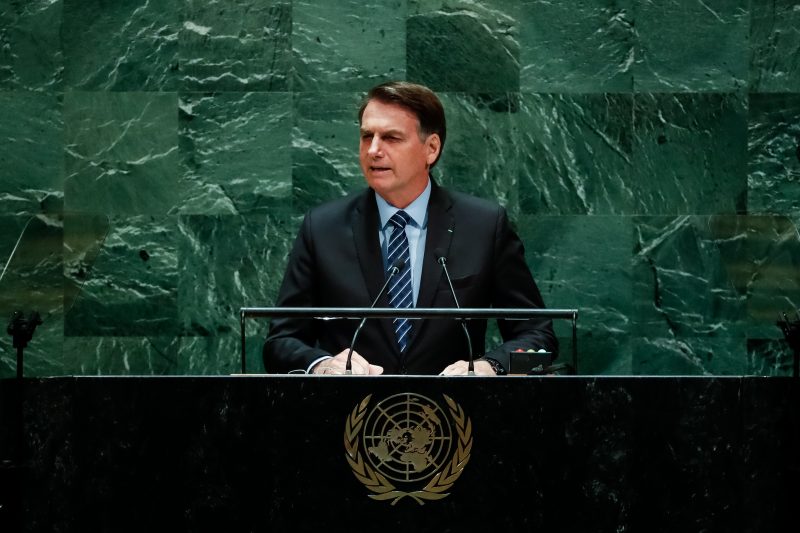 Presidente Bolsonaro discursando e usando terno preto e o fundo com piso verde escuro e na frente é possível ver o o símbolo da ONU