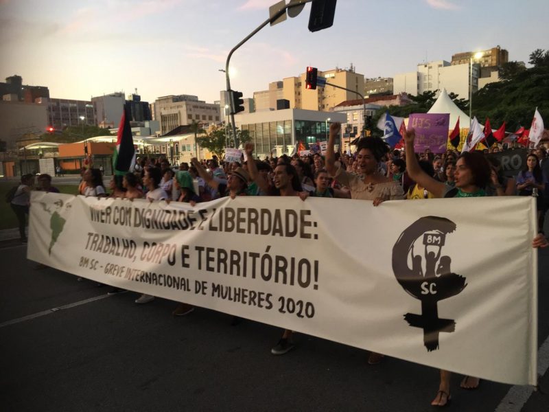 Foto mostra mulheres na rua segurando bandeiras durante a marcha do Dia Internacional da Mulher
