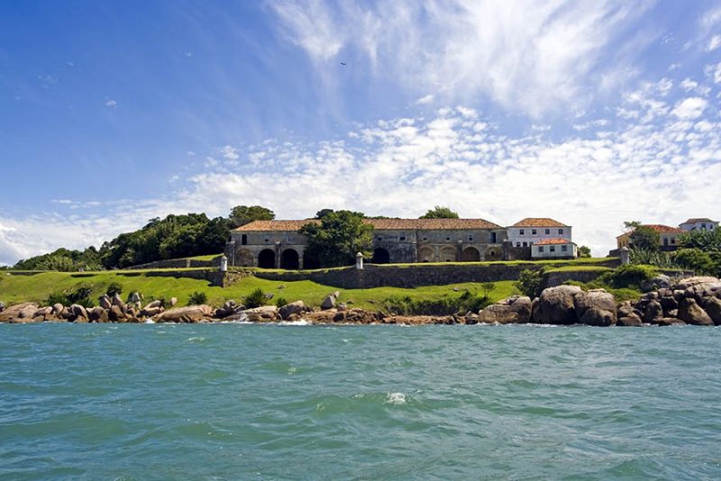 Foto da Fortaleza de Anhatomirim, cima de um pequeno morro verde e cercada por pedras. À frente dela está o mar. 