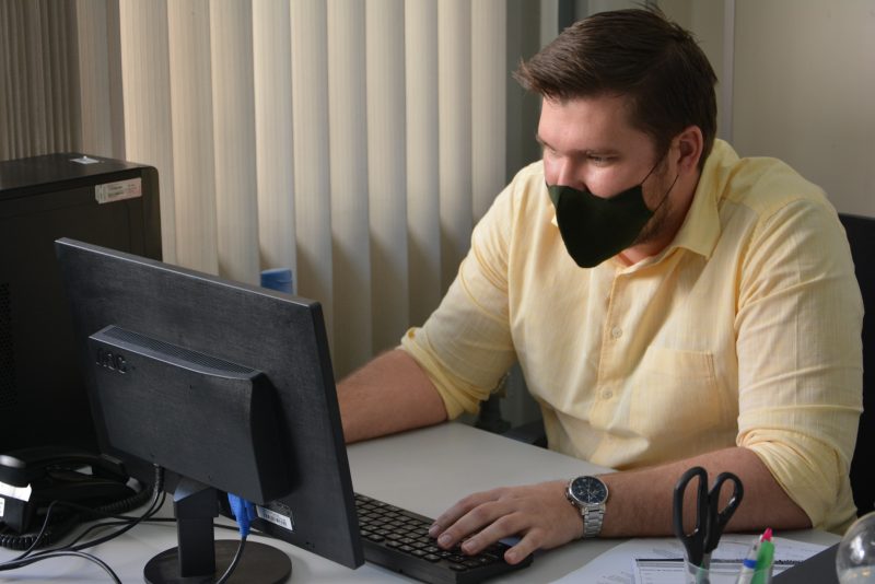 Homem usando máscara de camisa amarela sentado à frente do computador
