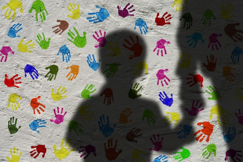Imagem da sombra de uma criança de mãos dadas com um adulto. Ao fundo uma parede braca com várias marcas de mãos de crianças coloridas