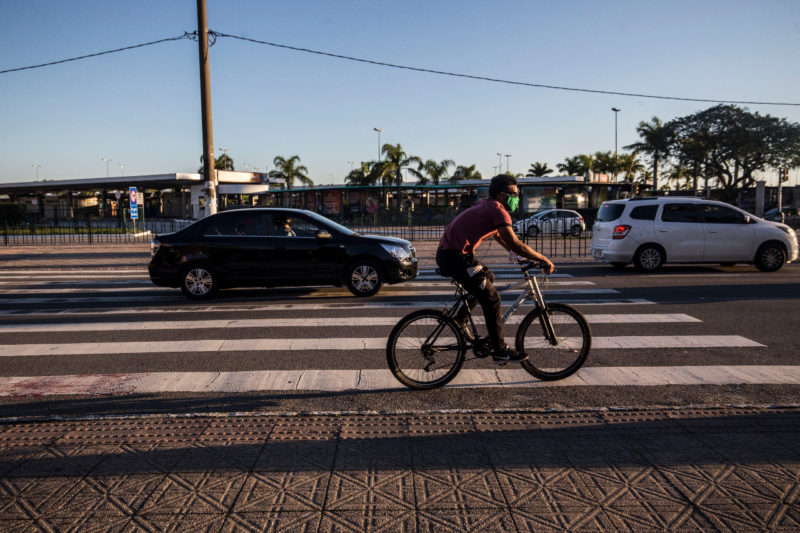 Pessoa andando de bicicleta e dois carros ao fundo, sendo um branco e outro preto passando pela faixa de pedestre. O céu está sem nuvens e ensolarado