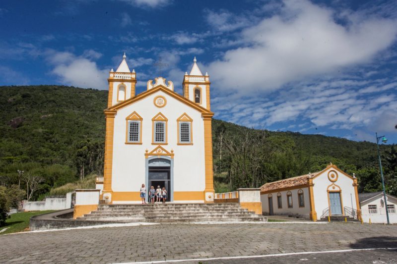 Foto da Igreja Nossa Senhora da Lapa, no bairro histórico de Ribeirão da Ilha, em Floripa. A igreja é pequena, com duas torres, parede branca e contornos amarelos. Ao fundo, um morro verde e um céu azul. 