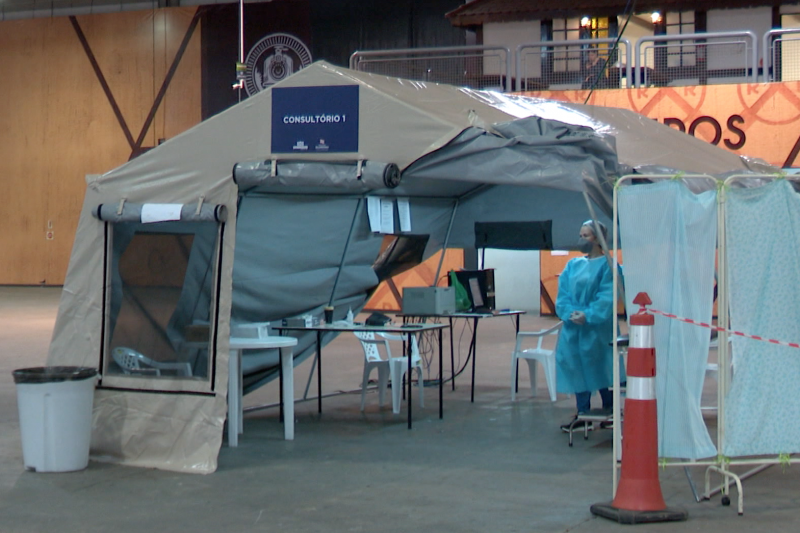 Imagem mostra uma tenda equipada com computador e equipamentos de atendimento médico. Na entrada há uma placa onde se lê "Consultório 1". Uma pessoa está parada na entrada da tenda. Ela veste touca, máscara e uma roupa azul de proteção para atendimento médico