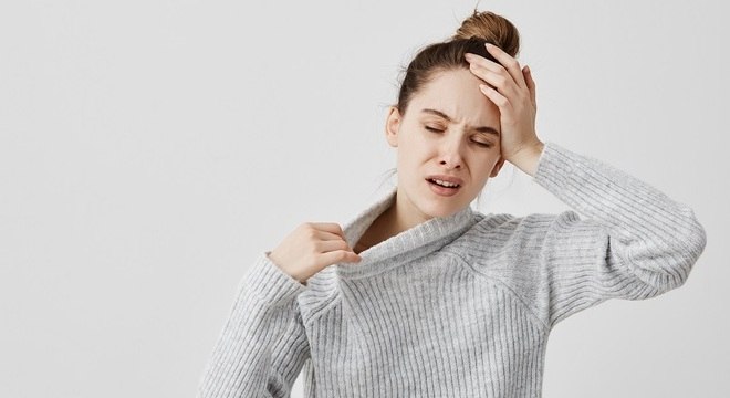 mulher com uma mão na cabeça e a outra puxando a gola da blusa com expressão de dor