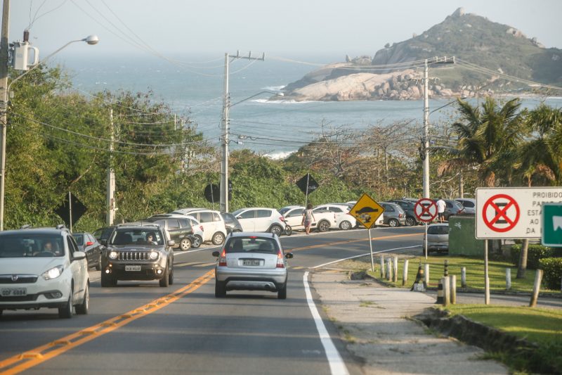 Trânsito mostra carros e mar ao fundo. Vários carros estacionados.