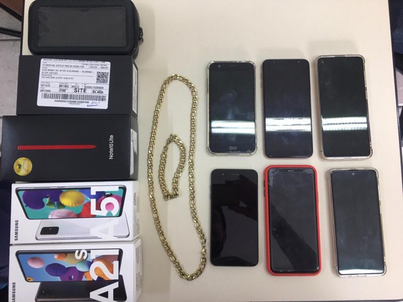 Foto do material apreendido pela polícia. São seis celulares e uma corrente de ouro. Tudo em uma mesa da delegacia. 