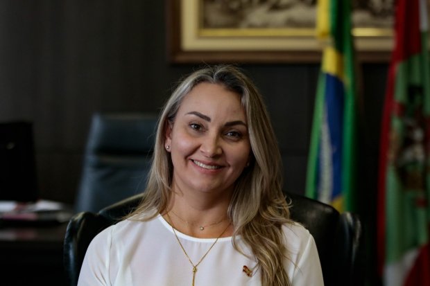 fotografia da governadora Daniela Reinehr. Ela está sentada, veste uma blusa branca, está com os cabelos soltos. Ao fundo aparecem as bandeiras de Santa Catarina e do Brasil.