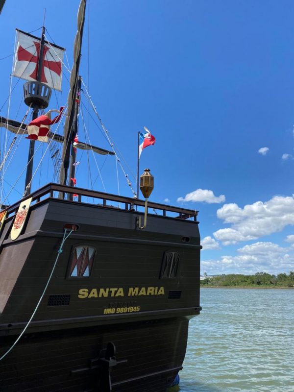 Embarcação NAU Santa Maria está no mar. O barco é marrom com dourado e bandeiras vermelhas. O céu está limpo
