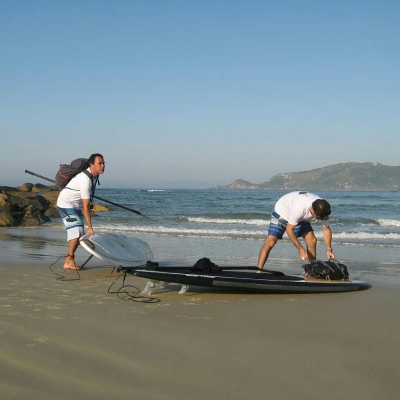 Dois homens estão na praia com duas pranchas de stand up paddle