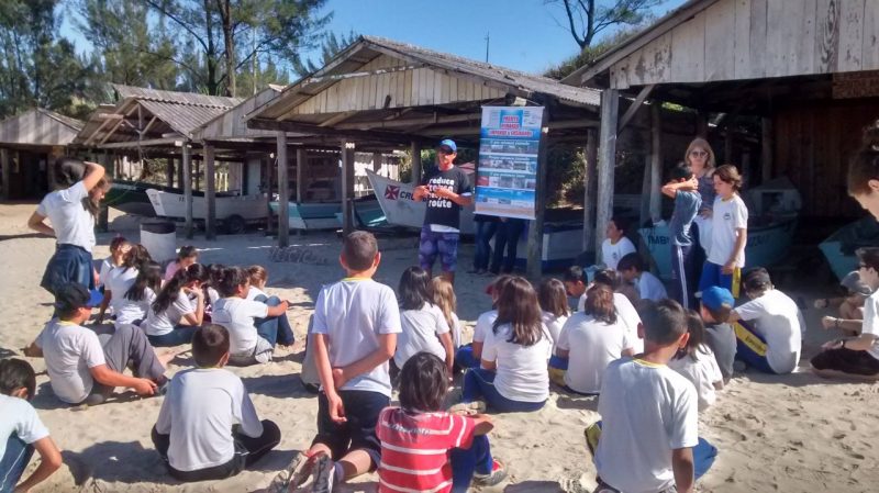 Crianças estão na praia sentadas enquanto assistem uma aula de educação ambiental