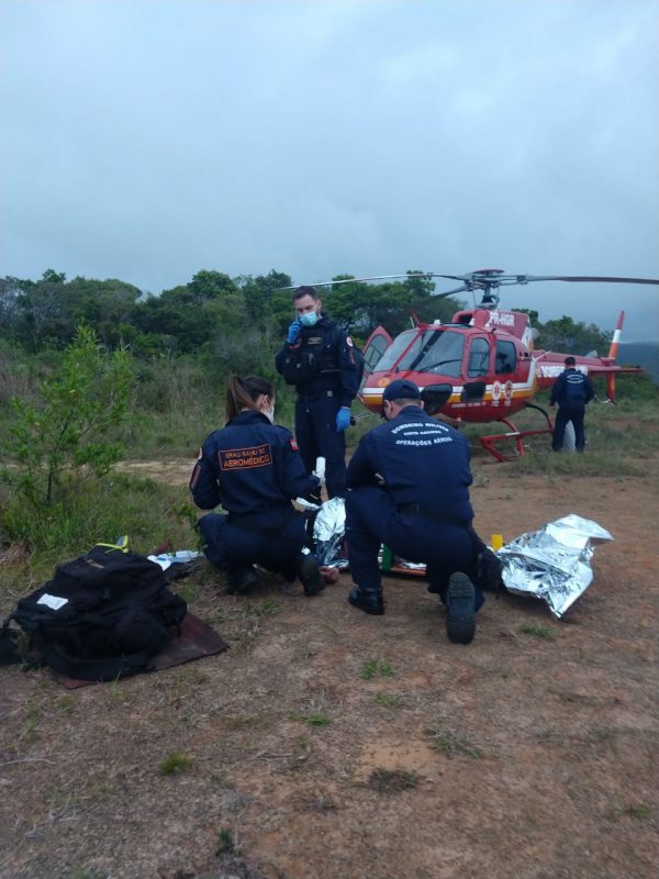 Dois bombeiros prestam atendimento a homem deitado. Ao fundo, helicóptero do Arcanjo.