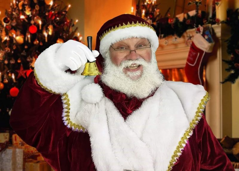 Papai Noel olha para a câmera e sorri. Ele segura um sino