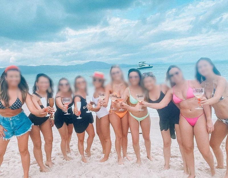 Dez mulheres posam para foto na areia. Mar ao fundo