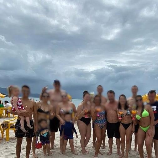 Grupo de banhistas entre crianças e adultos posam para foto na areia e mar ao fundo.