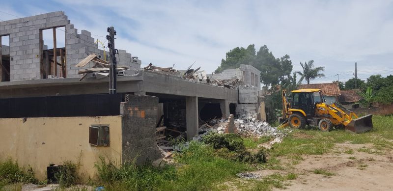 Construções irregulares sendo demolidas. Aparece uma construção pela metade e mato. 