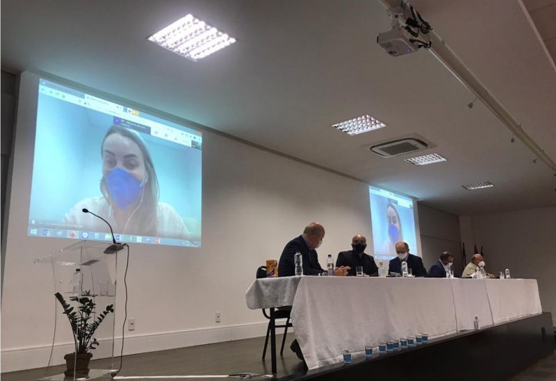 Fotografia de uma mesa com várias autoridades sentadas. Ao fundo tem um telão onde aparece a governadora de Santa Catarina. É uma imagem da reunião.
