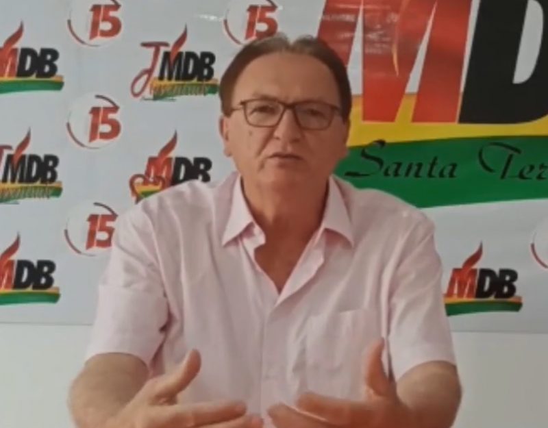 Fotografia do candidato Genir. Ele veste camisa de manga curta na cor rosa e usa óculos de armação vermelha. Ao fundo da imagem tem logomarcas do partido do candidato, o MDB