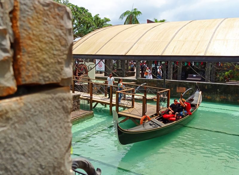 Foto mostra um pequeno barco, uma gôndola italiana, dentro de um lago artificial. Há um casal dentro dela. Ao lado há uma passarela de madeira que dá acesso ao barco. A atração fica na cidade de Nova Veneza, em Santa Catarina, um bom local para passar a lua de mel. 