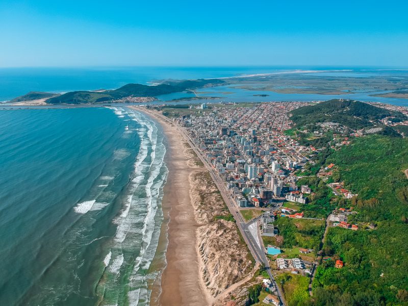 Foto da Praia do Mar Grosso, em Laguna. No canto esquerdo da imagem, o mar. No canto direito, a faixa de areia, construções urbanas e áreas verdes. 