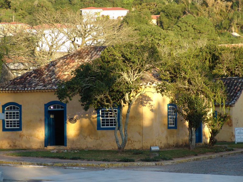 Foto do Museu Casa de Anita, um casarão amarelo com janelas azuis. à frente dele há uma árvore. Atrás, aparecem algumas construções cercadas pela vegetação. 