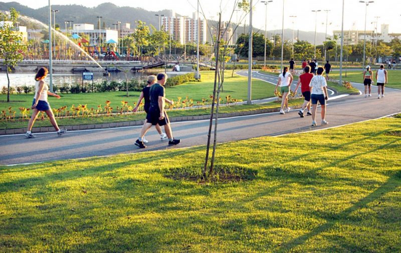 Foto de pessoas caminhando em um parque. Ao fundo é possível observar um lago e o parque infantil.