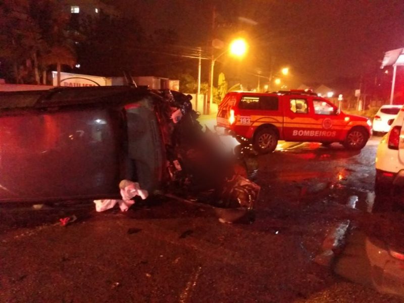 Foto mostra cena de acidente de trânsito. Um carro está capotado de lado, com o teto do carro em primeiro plano. É possível ver também um air bag acionado. Ao fundo há uma viatura do Corpo de Bombeiros. A imagem está escura porque foi registrada à noite. A cena é iluminada pela luz de um poste.