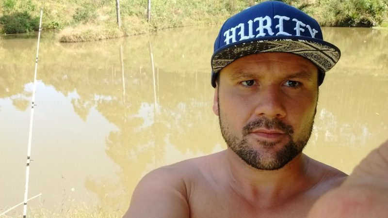 Foto do homem assassinado. Ele está sem camiseta. Usa um boné. Aparenta estar escando, pois tem varas e um lago ao fundo da imagem. 