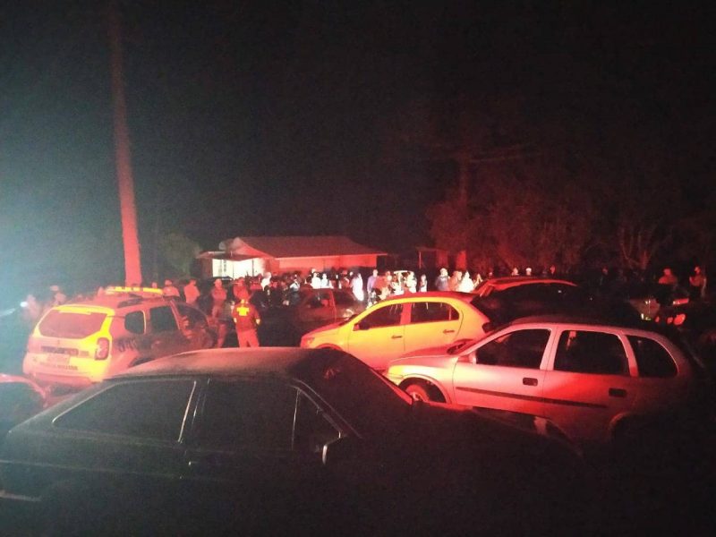 Foto escurada onde aos fundos tem uma casa. Várias pessoas estão do lado de fora, com muitos carros e aparece também a viatura da Polícia Militar. 