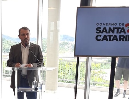 Fotografia do governador falando em um púlpito. Ao lado dele há uma tv com a bandeira do governo do estado de santa catarina.