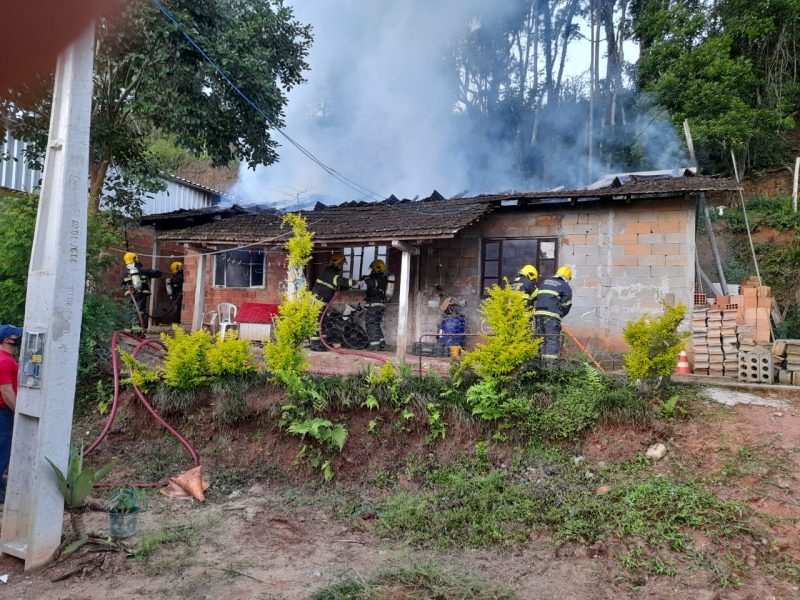 Casa de tijolos parcialmente rebocada com vários bombeiros combatendo um incêndio. É possível ver fumaça saindo pelo telhado.