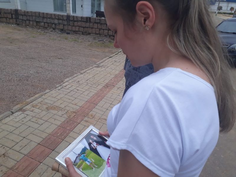 Foto de uma mulher loira de lado. Ela está de cabeça baixa, com olhar triste, observando um porta-retrato com fotos do irmão.
