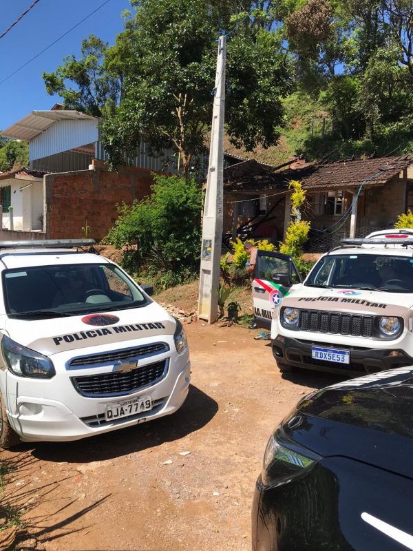 Foto de uma rua de barro com duas viaturas da pol´ciia militar. Aos fundos aparecem algumas casas. 