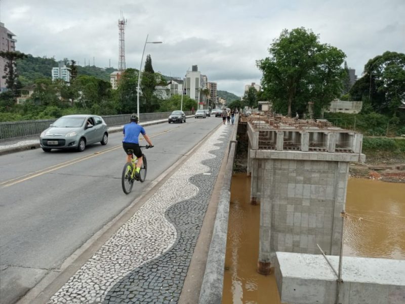 Foto da Ponte Adolfo Konder no Centro de Blumenau, com carros trafegando e um ciclista. Ao lado é possível ver o andamento da obra de duplicação da estrutura.