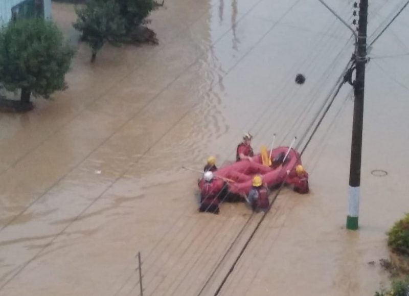 Imagem de uma rua alagada e bombeiros com um bote. 