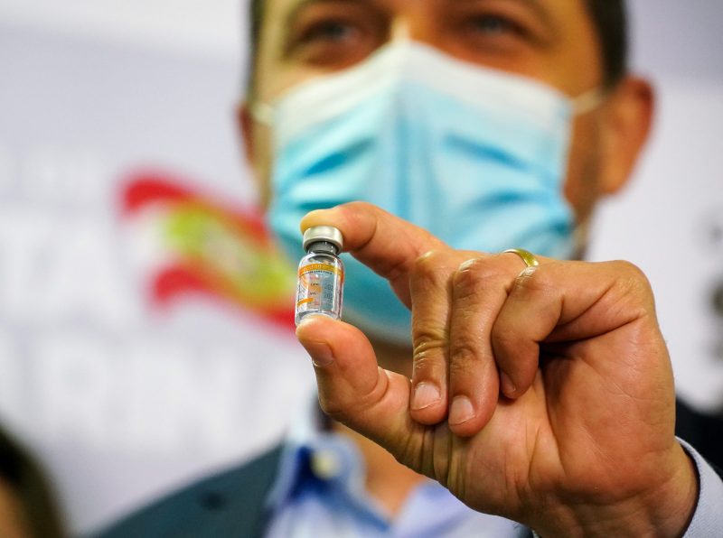 Governador Carlos Moises segurando uma das doses da vacina contra a Covid-19, na imagem ele está de máscara azul e o seu rosto está desfocado com foco da foto na mão dele segurando a vacina, no seu dedo tem uma aliança de ouro