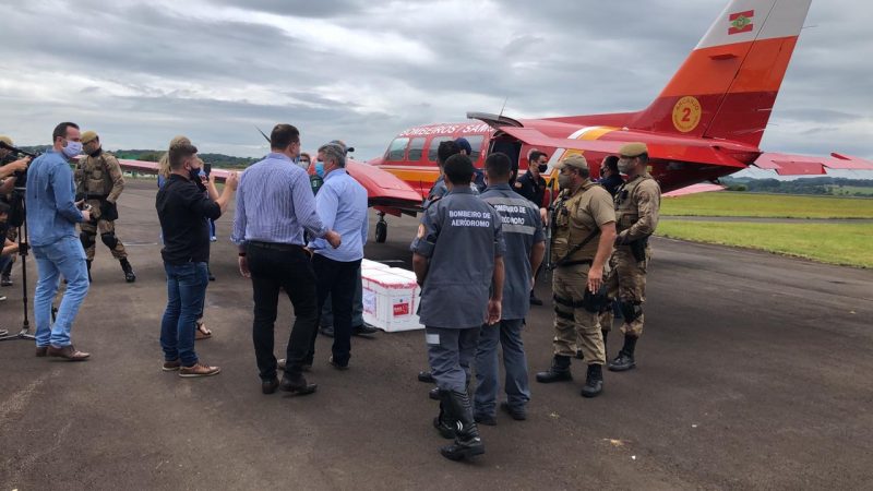 Aeronave arcanjo do corpo de bombeiros pousado no aeroporto de chapecó, a aeronave é vermelha com as faixas amarela e branca e o símbolo da bandeira de SC, Na pista várias pessoas se aproximam do avião, profissionais da imprensa, bombeiros e policiais militares, todos de máscara. As caixas com as vacinas contra a covid-19 são brancas de isopor com informações em vermelho