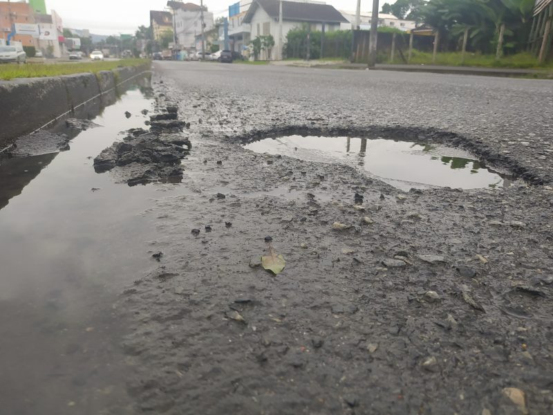 Foto de uma rua asfaltada em que aparece um buraco cheio de água no meio da pista.