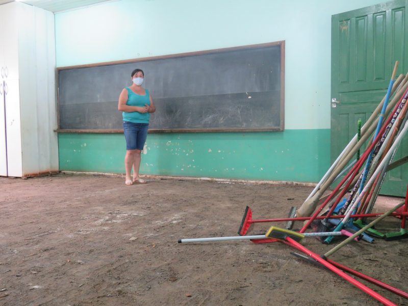 Foto de uma sala de aula. Uma mulher está em frente ao quadro e o chão está sujo de terra. No canto direito há materiais de limpeza, como rodo, vassouras e escovão.