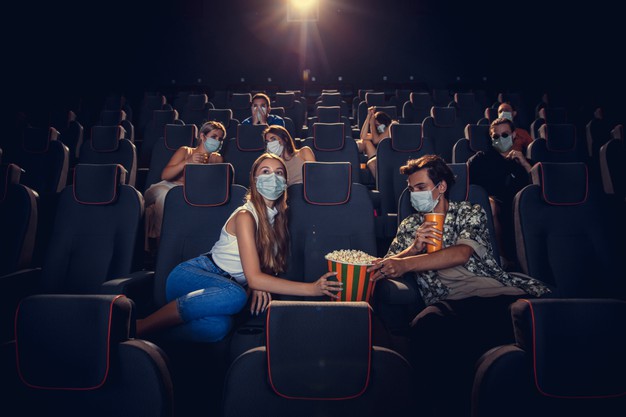 Sala de cinema com pessoas sentadas nas cadeiras e usando máscara, elas estão em distanciamento social.
