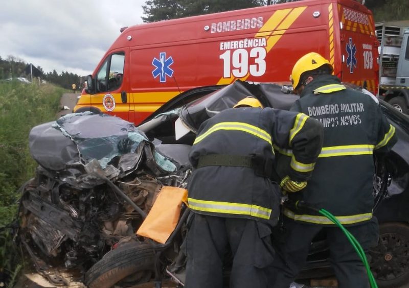 Na imagem aparecem dois bombeiros militares atendendo a vítima que está presa às ferragens do carro pálio. O carro está todo danificado e amassado. Ao fundo tem a ambulância do corpo de bombeiros. O jovem morreu no local do acidente em Campos Novos.