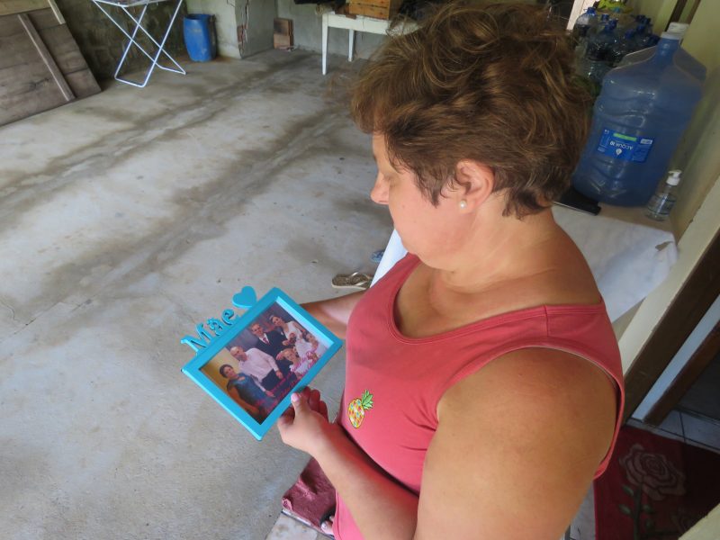 Foto de cima de uma mulher de cabelo curto olhando para um porta retrato escrito mãe. Nela há uma foto dela com o marido o filho e a nora.