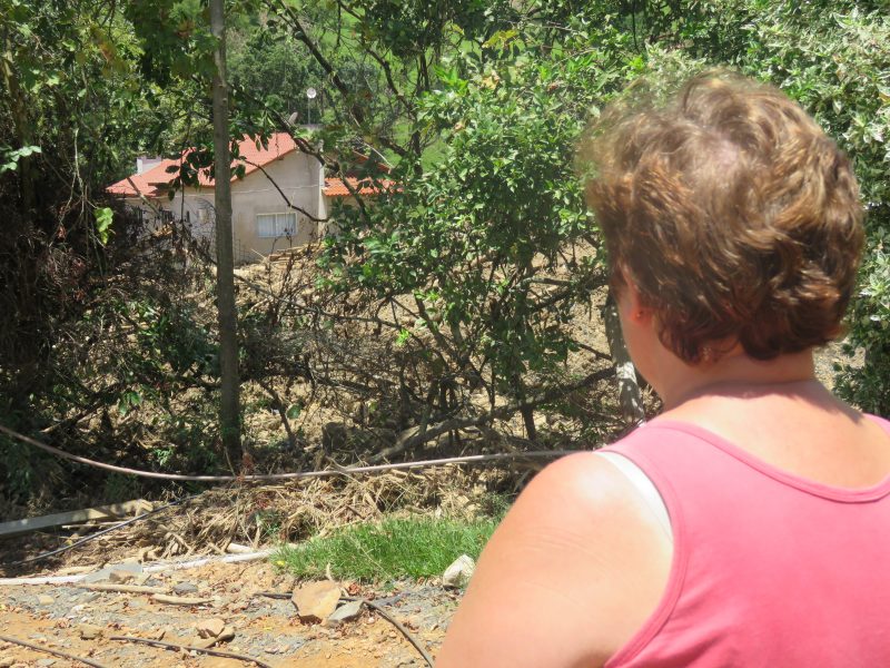 Foto de uma mulher de cabelo curto e blusa rosa. Ela está de costas olhando para frente. Ao fundo é possível ver parte de uma casa de material sem pintura.