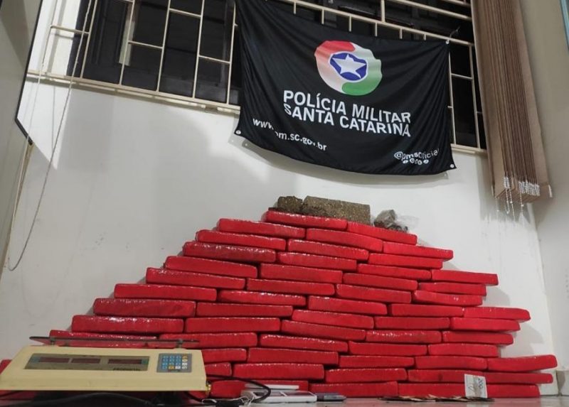 Foto de uma sala branca com janela gradeada. Vários tabletes de drogas embrulhados em pacotes vermelhos estão empilhados.