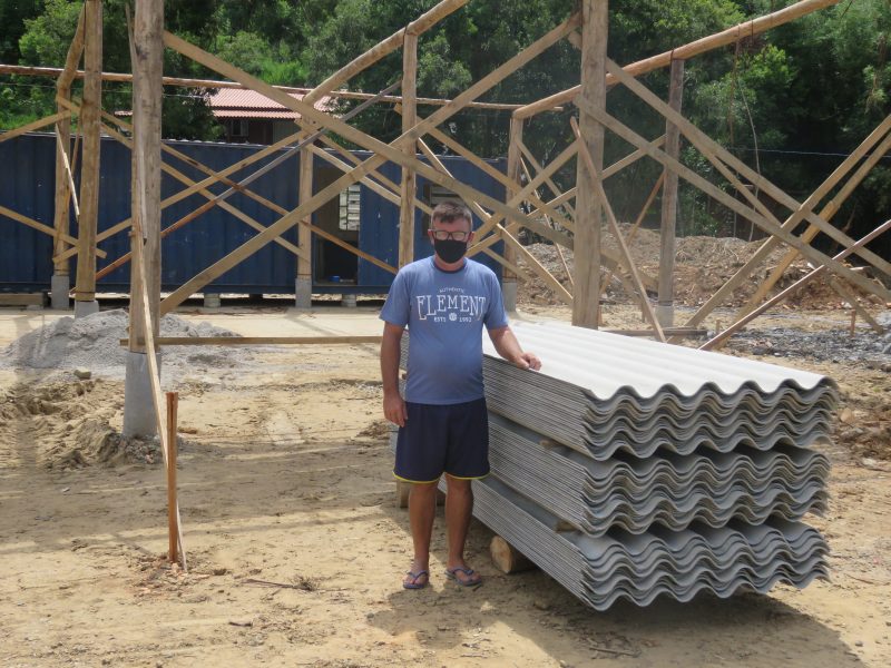 Um homem de máscara e óculos está lado de um monte de eternit e ao fundo há uma obra.